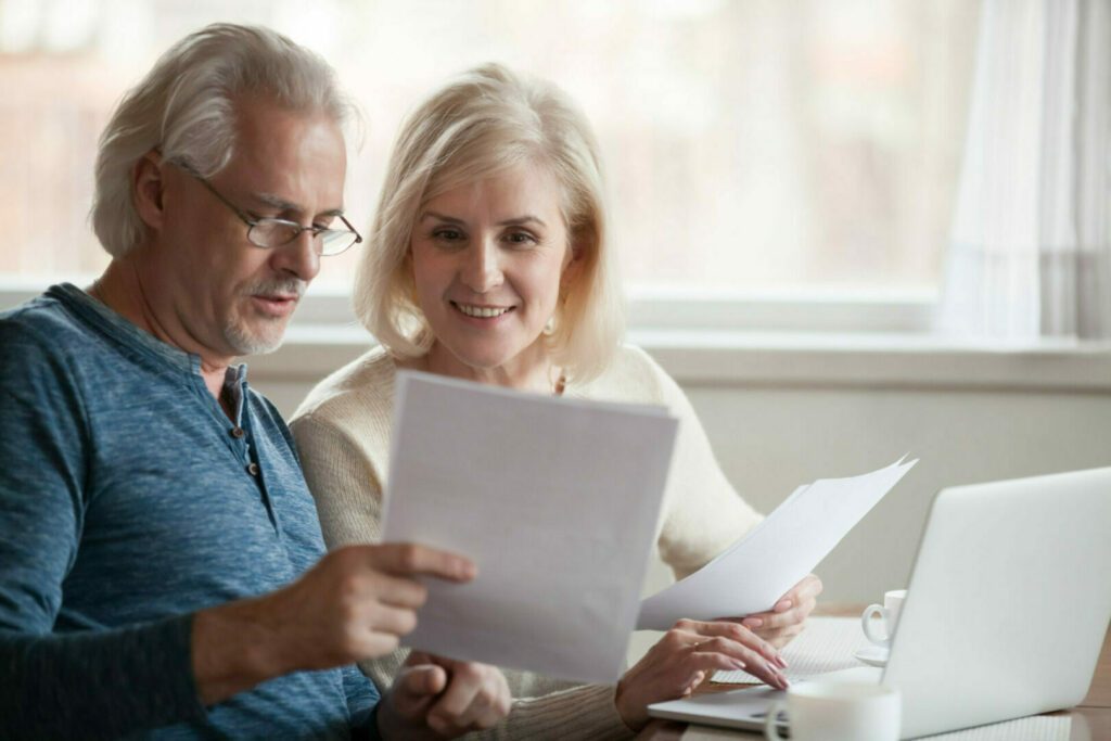 Happy old middle aged couple holding reading good news in document, smiling senior mature family excited by mail letter, checking paying domestic bills online on laptop, discussing budget planning